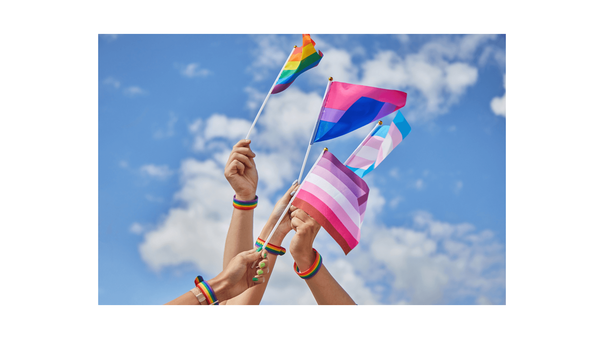 Four hands wearing rainbow bracelets hold up various LGBTQIA+ flags against a blue sky.